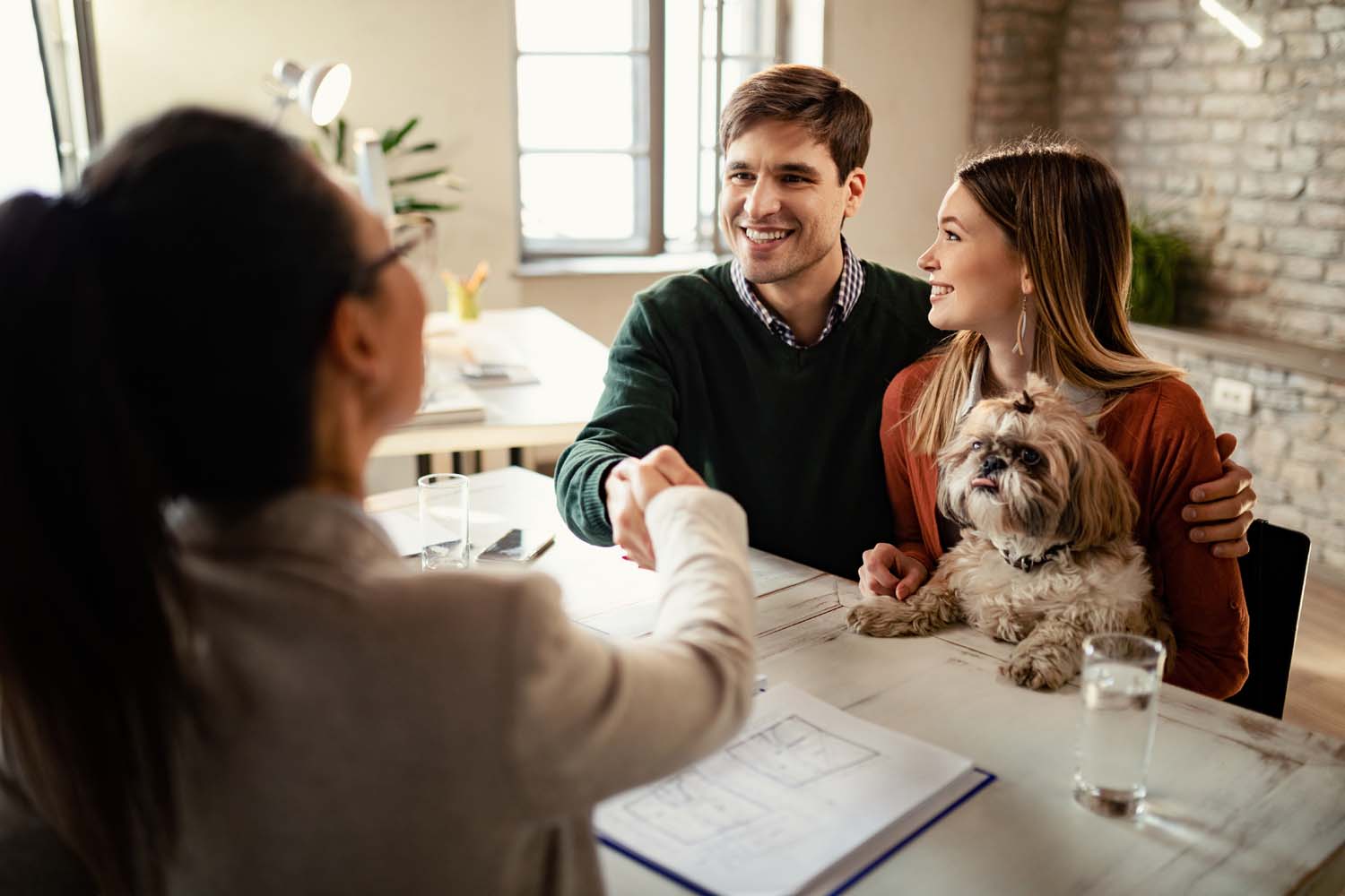 Happy couple with a dog shaking hands with real estate agent in the office.